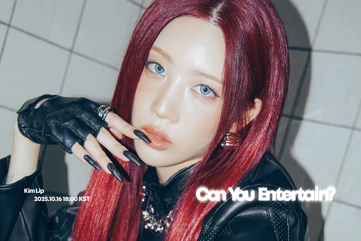 A close-up portrait of a young woman with long, red hair and striking blue eyes, wearing a black leather outfit and fingerless gloves, posed against a tiled background. Text in the image reads 'Can You Entertain?' and includes a date and time.