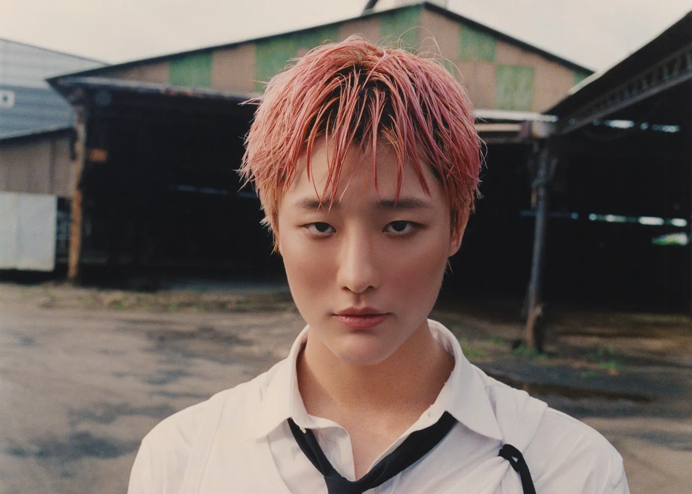A close-up portrait of a person with short, wet pink hair, wearing a white shirt and black tie, standing outdoors with a blurred industrial background.