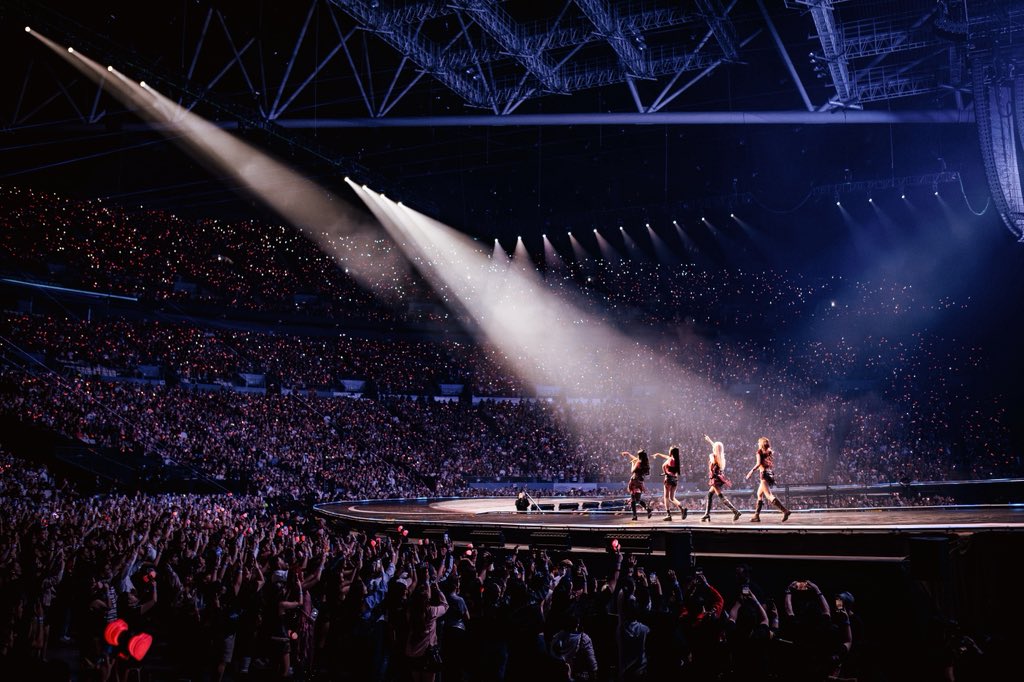 A vibrant concert scene featuring BLACKPINK performing on stage with a large, enthusiastic crowd in the foreground, surrounded by dramatic lighting and colorful glow sticks. BLACKPINK concert DEADLINE World Tour 2025 at Philippine Arena, Bulacan, Philippines