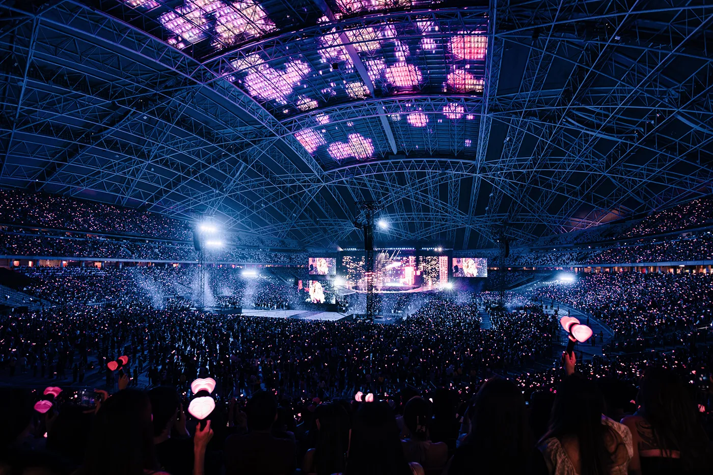PHOTO BLACKPINK DEADLINE SINGAPORE 2025: A large crowd at a BLACKPINK concert in a stadium, illuminated by colorful lights, with fans holding pink light sticks.