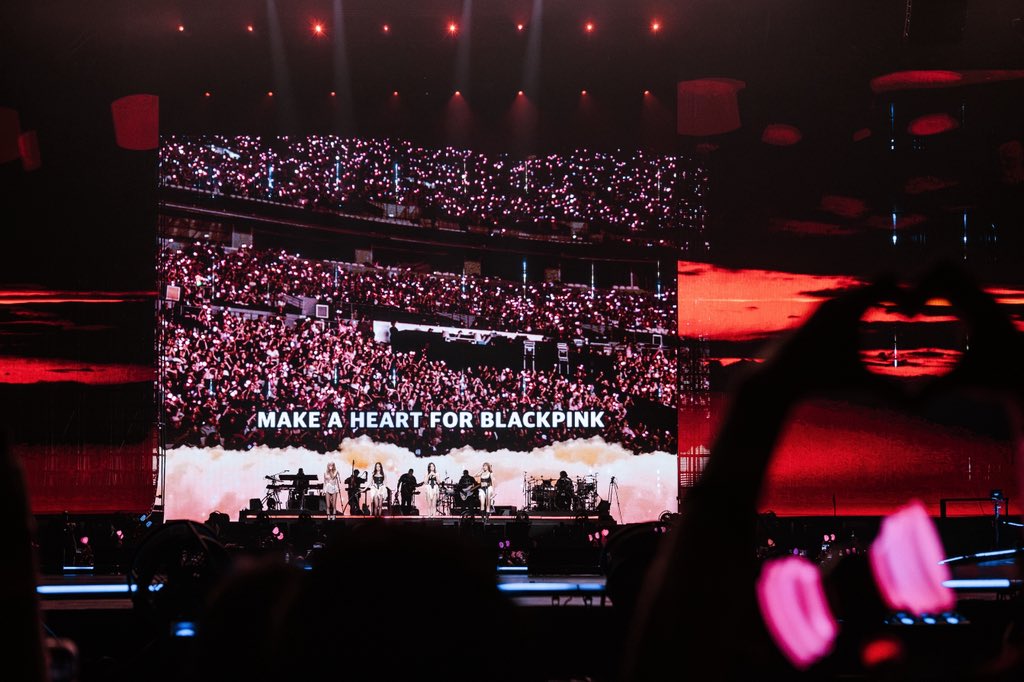 A vibrant concert scene featuring the audience holding lightsticks, with a large screen displaying the text 'MAKE A HEART FOR BLACKPINK' during a BLACKPINK performance.