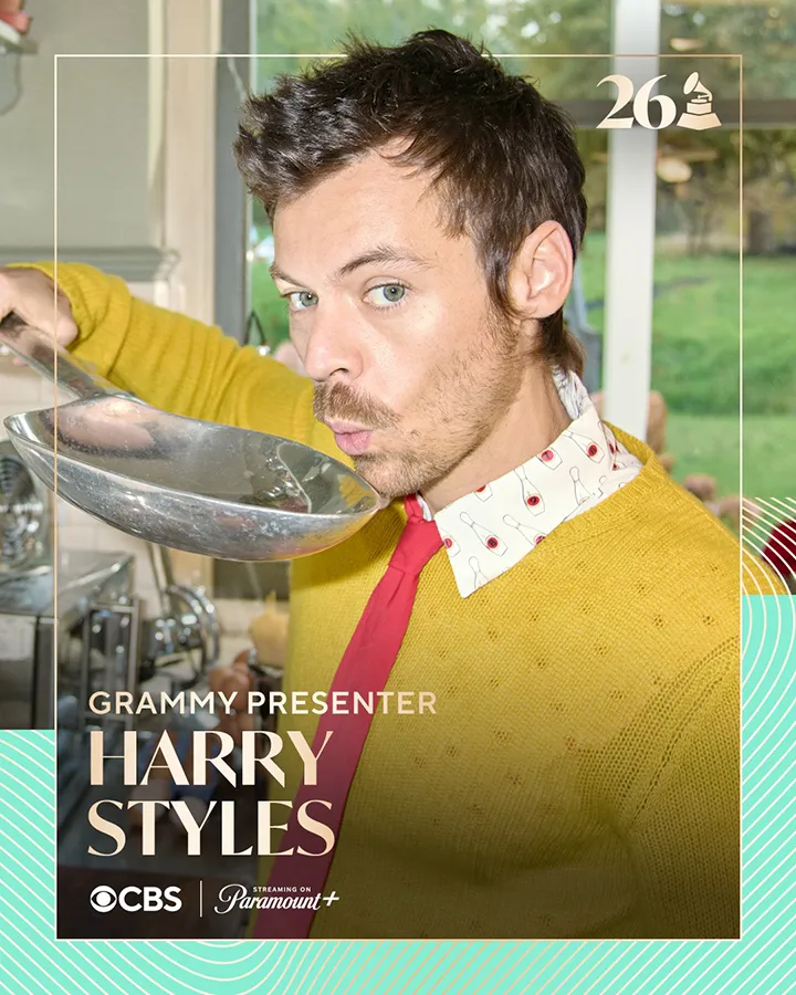 A man with short, dark hair is holding a large spoon and pretending to sip from it. He is wearing a bright yellow sweater over a red shirt with a patterned collar. The background features greenery and a kitchen setting, with a notation indicating he is a Grammy presenter.