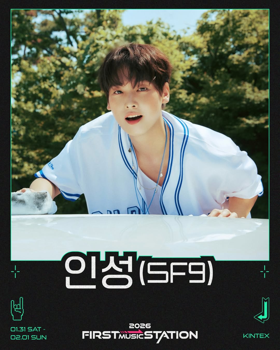 A young man in a white jersey with blue accents smiles while leaning over a car, surrounded by greenery.