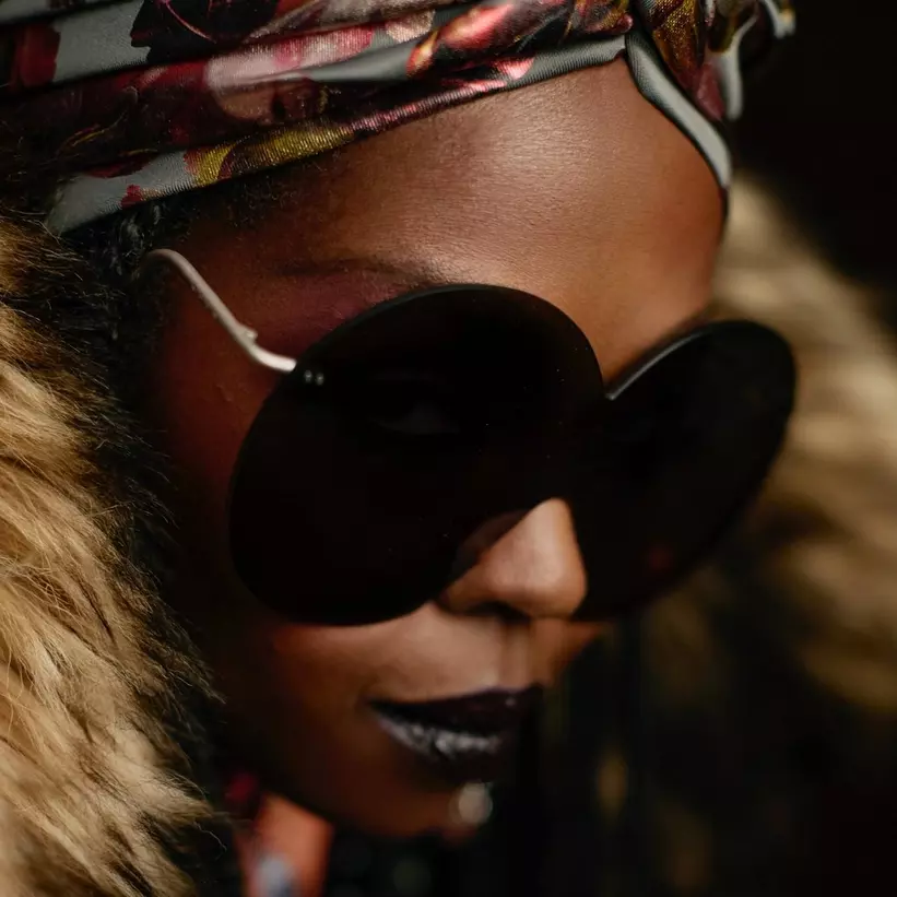 Close-up of a person wearing large black round sunglasses and a colorful headscarf, with fur accents around their collar.