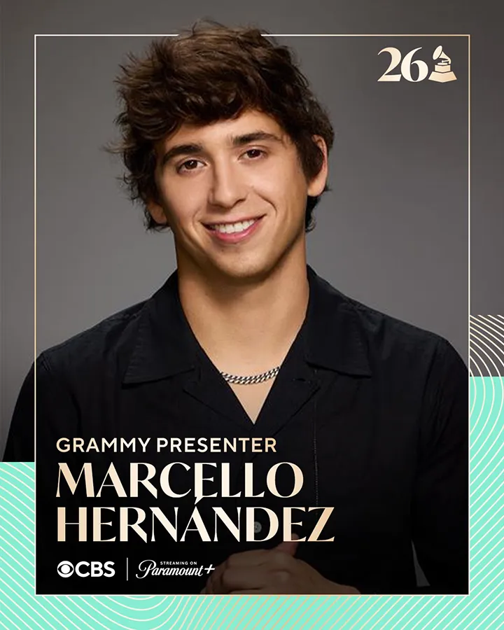 Portrait of Marcello Hernandez, a Grammy presenter, smiling and wearing a black shirt with a chain necklace, set against a dark background.