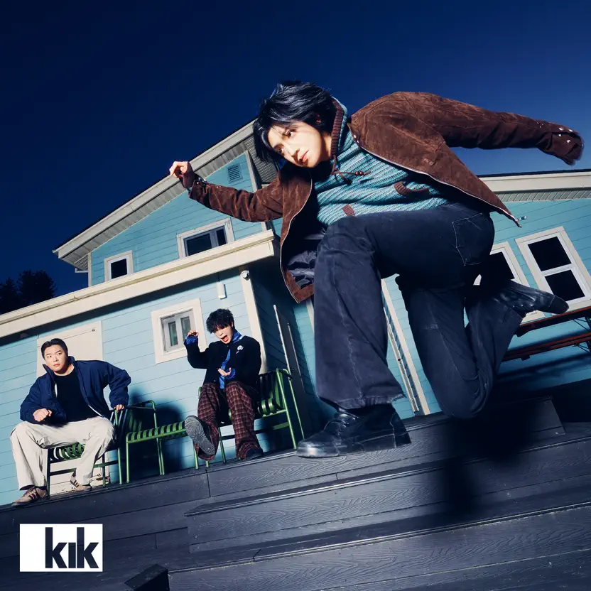 Three individuals pose in front of a blue house, with one jumping energetically in the foreground, while the others watch. The scene is set against a dusk sky.