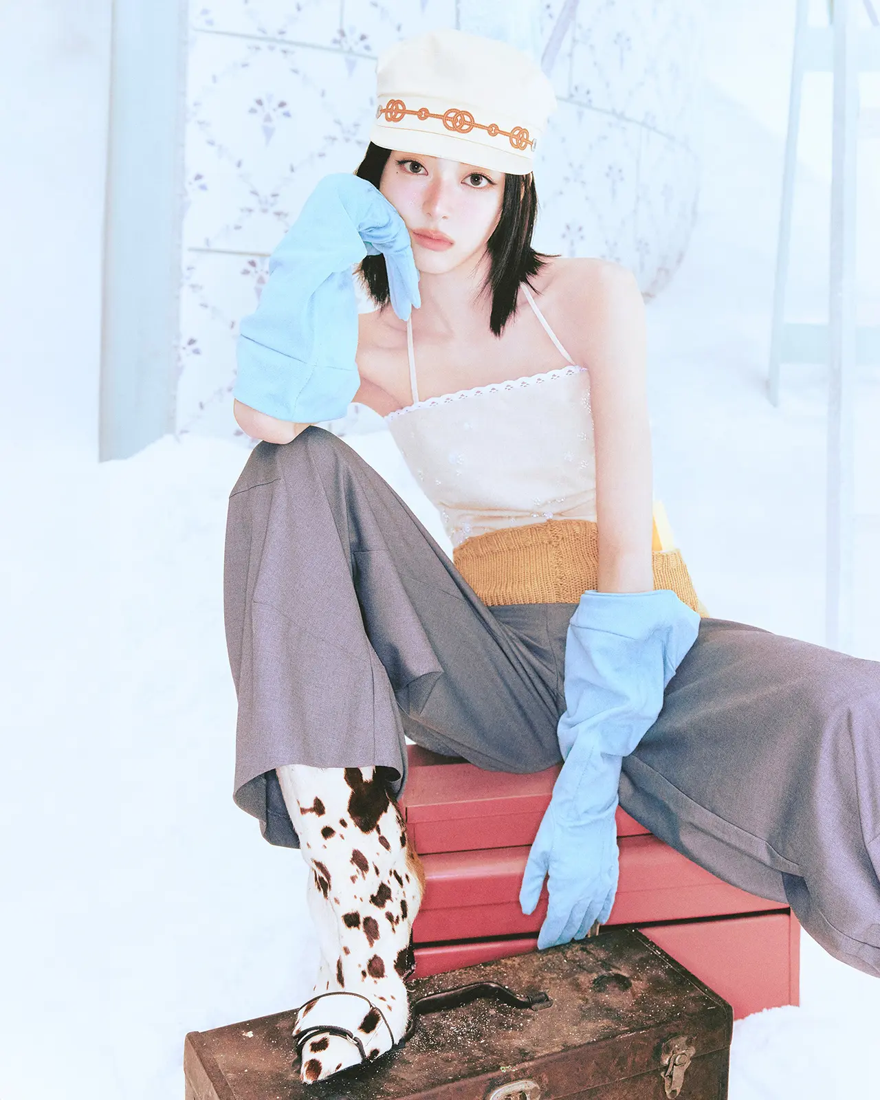 ITZY Yuna for 1st Mini Album Ice Cream. Yuna sitting on a red chest, wearing a cream cap, a light top, and gray pants, with blue rubber gloves and cow-print boots, set against a snowy background.