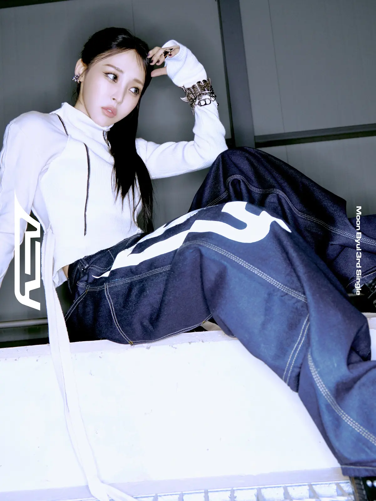 Moon Byul Teaser for 3rd Single REV. Moon Byul sitting casually in a modern outfit, featuring a white long-sleeve top and dark oversized pants, with accessories including rings and bracelets. The setting has an urban aesthetic.