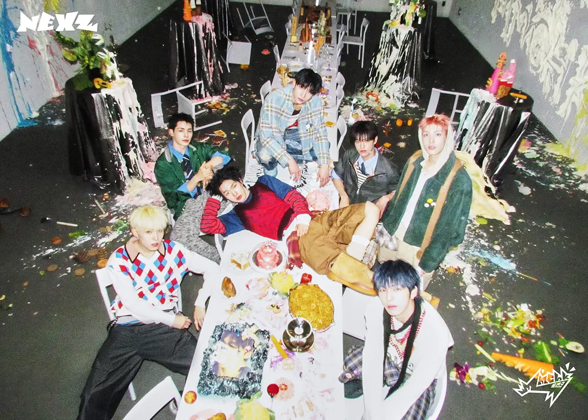 Teaser for NEXZ 2nd Single Album – Mmchk (2026). NEXZ members sitting around a messy dining table filled with colorful food and decorations, surrounded by a chaotic backdrop with paint splatters and party decor.