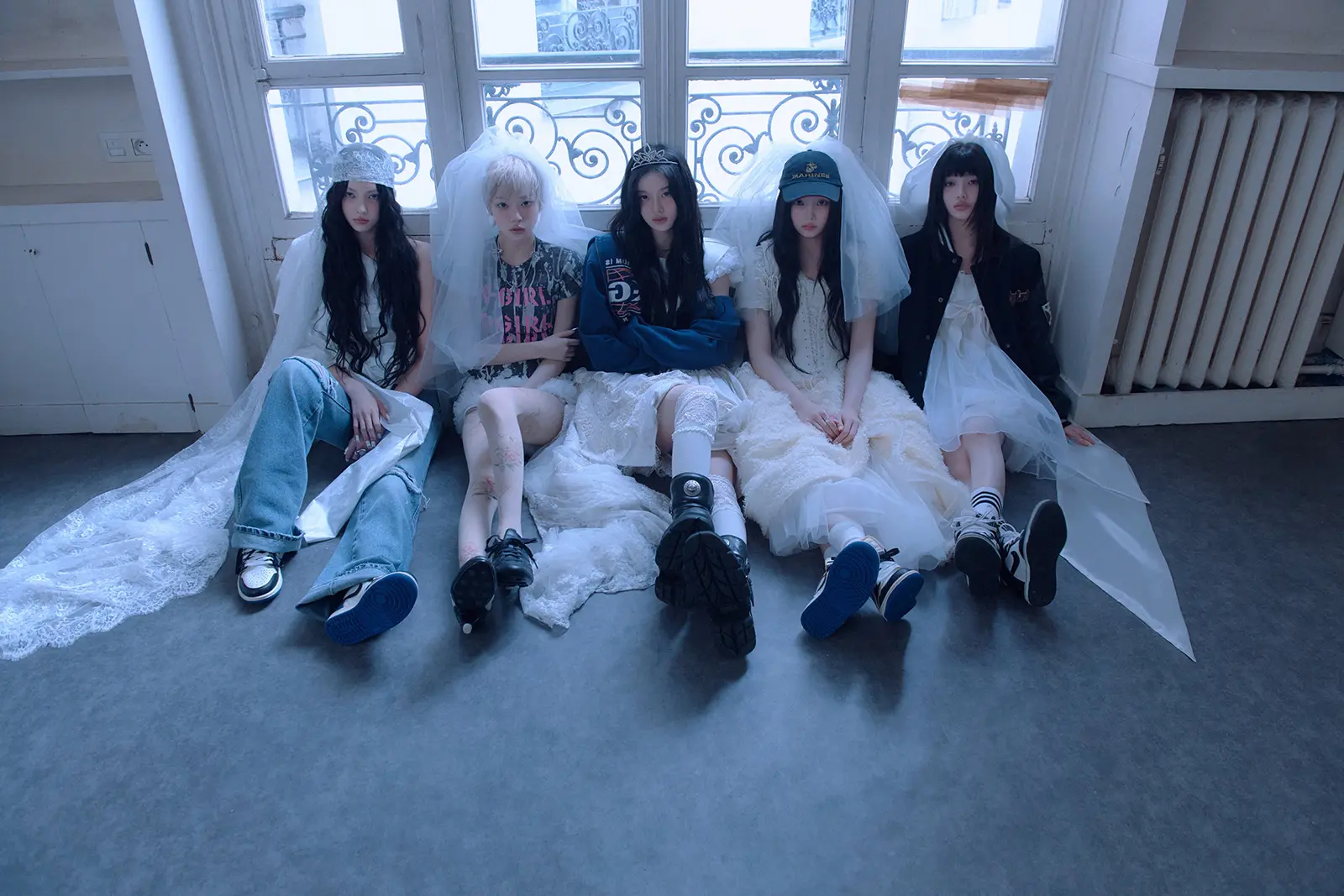 Teaser for ILLIT 4th Mini Album – MAMIHLAPINATAPAI (2026). Five individuals sitting on a couch, wearing a mix of casual and elegant attire, including veils and lace. They are positioned in front of large windows with decorative ironwork.