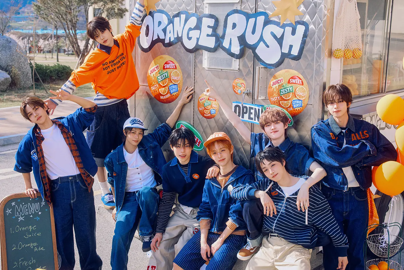 Teaser for YUHZ 1st Single Album – Orange Record (2026). Group of eight young people posing in front of a colorful food truck named 'Orange Rush', featuring various fruit-themed decorations and a menu board.