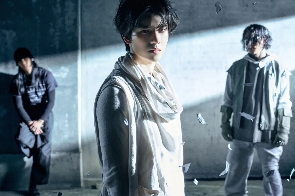 YORCH (POW) 1st Single – Blow Your Mind (2026). Three individuals pose dramatically in a dimly lit industrial setting, with light casting strong shadows. The foreground features a person in a white outfit, while two others in darker clothing stand behind, surrounded by scattered, floating debris.