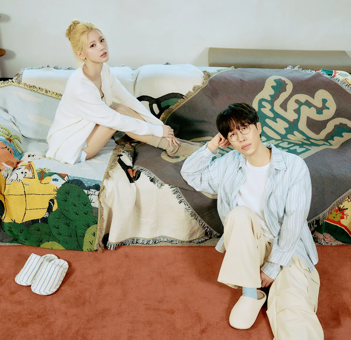 SORAN and MIYEON of i-dle. A young woman in a white sweater sits on a couch with colorful blankets, while a young man wearing glasses and a striped shirt leans against the couch, looking thoughtful. Soft textures and a cozy atmosphere enhance the scene.