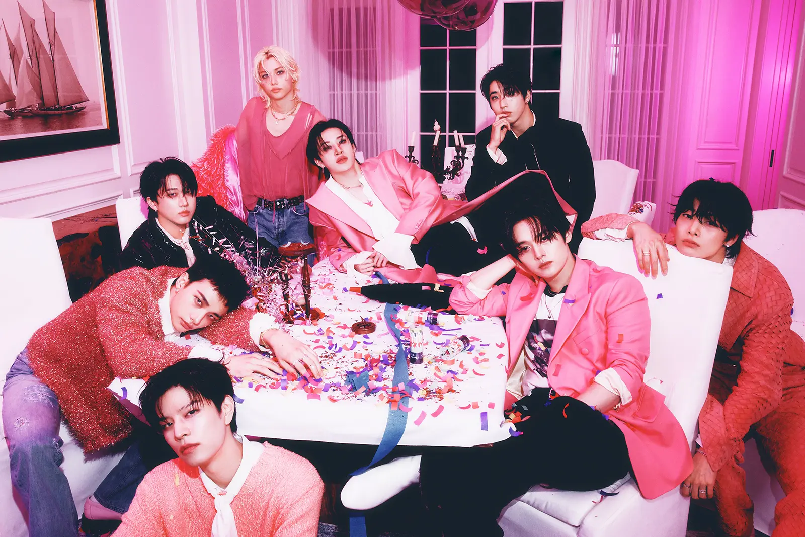 Stray Kids Members for SKZ IT TAPE EP – DO IT (2025). A group of nine people posed around a decorated table with colorful confetti. They are dressed in vibrant, trendy outfits, with a pink and purple theme visible in the background and lighting.
