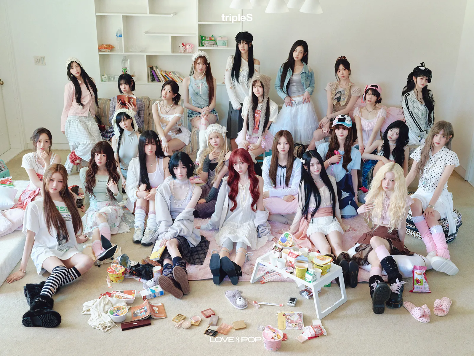 tripleS full members 3rd Album – ASSEMBLE26 LOVE&POP pt.1 (2026). A group of young women sitting together in a cozy indoor setting, dressed in various styles, surrounded by scattered snacks and playful accessories.