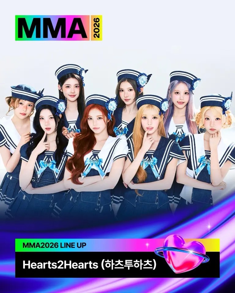 A group of eight female performers dressed in sailor-style outfits with blue accents, posing together for the MMA 2026 event under the name 'Hearts2Hearts (하츠투하츠).' The background is light-colored, with the 'MMA 2026' logo and a graphic featuring a heart.